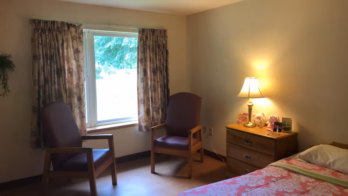A cozy bedroom with two wooden armchairs with purple cushions near a window with floral curtains. There is a wooden nightstand with a lit table lamp, some decorative items, and a framed photo. A bed with a pink floral bedspread is partially visible on the right side.