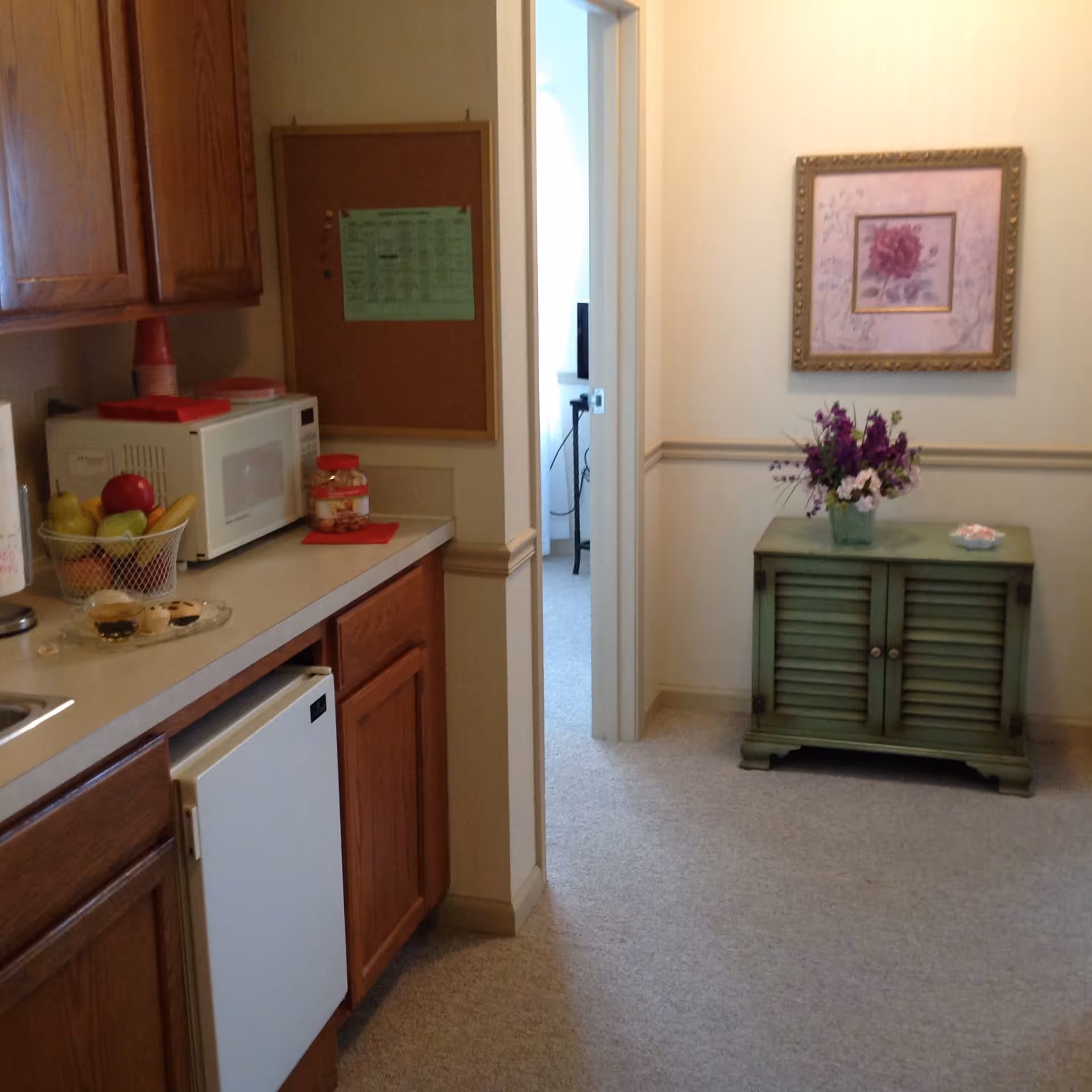Interior view of a small kitchenette area with wooden cabinets, a microwave, a mini refrigerator, and a countertop holding a fruit basket and snacks. Adjacent to the kitchenette is a hallway with a green cabinet topped with a vase of purple flowers and a framed floral picture on the wall.