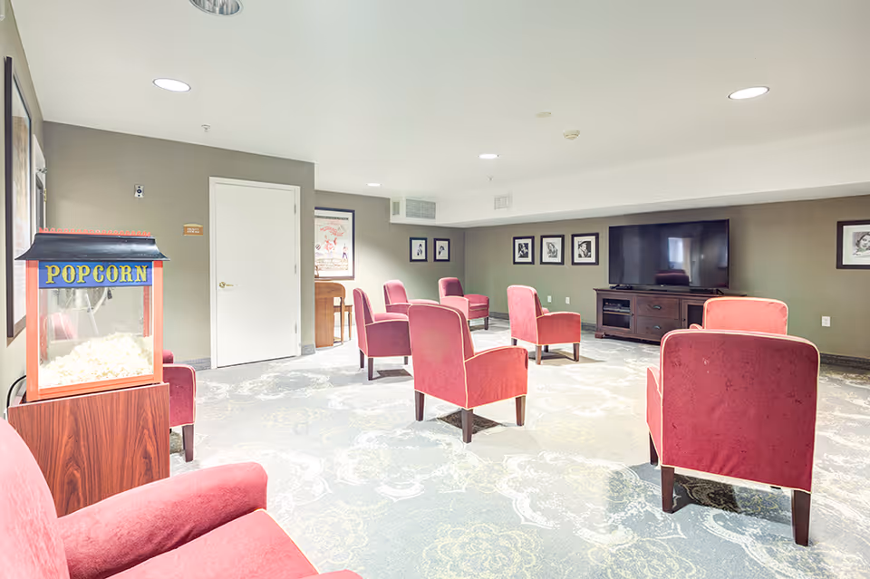 A senior living facility common room with several red upholstered armchairs arranged facing a large flat-screen TV on a wooden stand. There is a popcorn machine on a wooden stand to the left side of the room. The walls are decorated with framed pictures and the carpet has a floral pattern.