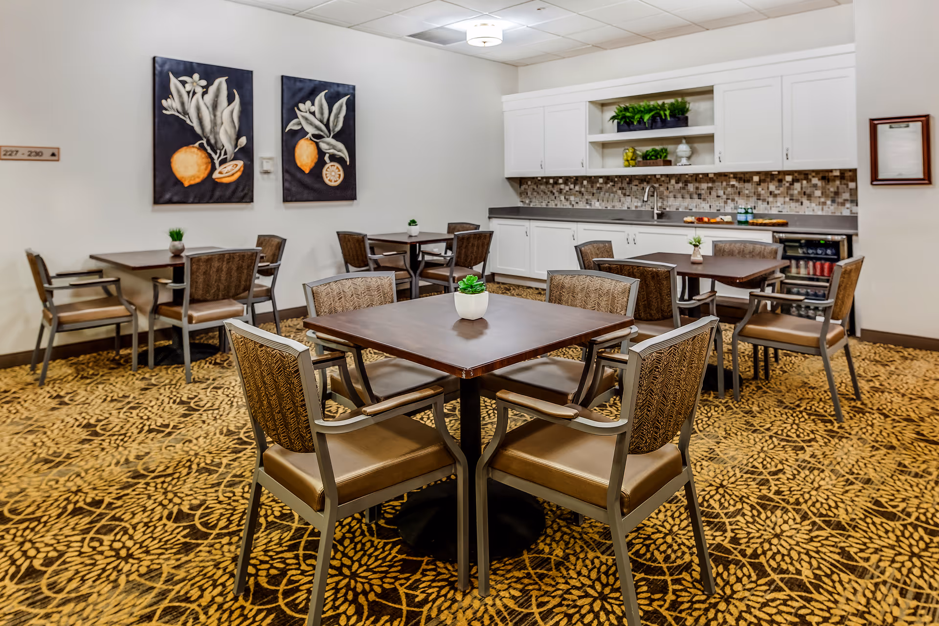 A dining area in a senior living facility with several square wooden tables, each surrounded by four cushioned chairs. The room has a patterned carpet and white walls adorned with two framed botanical prints. In the background, there is a kitchenette with white cabinets, a sink, a small refrigerator, and decorative plants on the countertop and shelves.