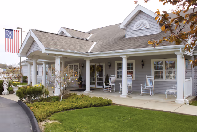 Exterior view of a single-story building with gray siding and white trim, featuring a covered porch with white columns and several white rocking chairs. An American flag is displayed on a pole near the porch. There is a well-maintained lawn and some shrubs in front of the building.