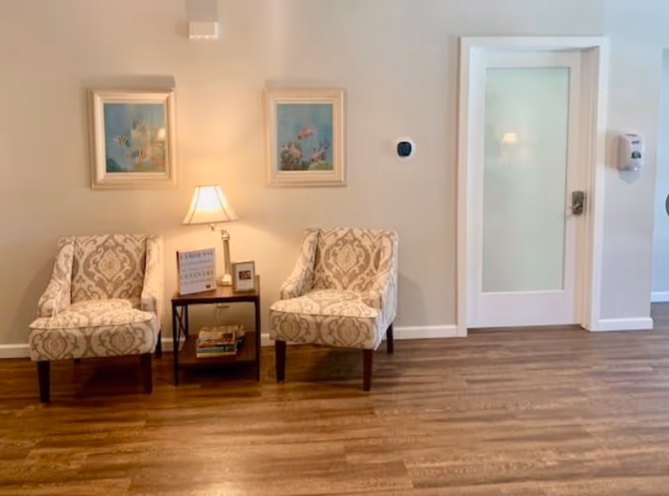 Two patterned armchairs with a small wooden side table between them holding a lamp, books, and a framed picture, set against a beige wall with two framed pictures hanging above. To the right is a white door with frosted glass and a wall-mounted hand sanitizer dispenser.