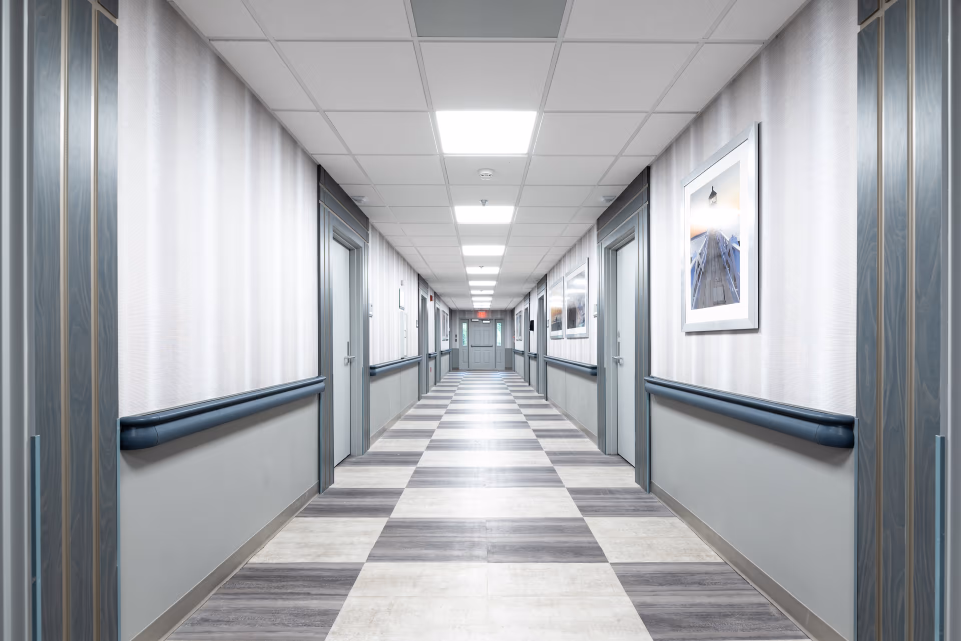 A long, well-lit hallway in a senior living facility with multiple closed doors on both sides, light-colored walls with framed pictures, and a patterned floor with alternating light and dark tiles.