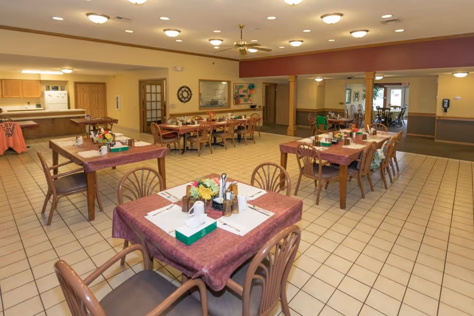 A spacious dining room in an assisted living facility with multiple tables covered in maroon tablecloths, each set with placemats, napkins, cups, and condiments. The room has tiled floors, beige walls, and ceiling lights. There is a kitchen area visible in the background and a bulletin board on one wall.
