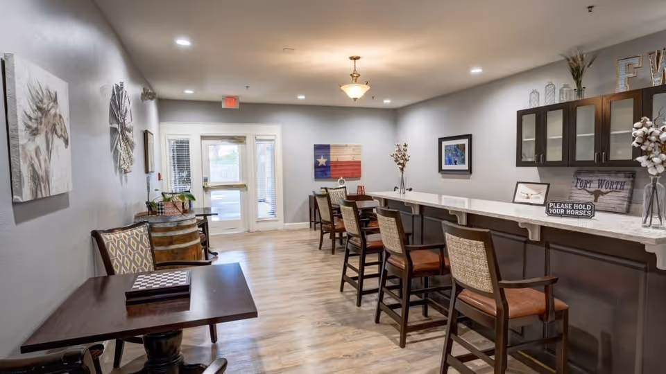 A well-lit interior common area with a long counter and several high chairs on the right side. On the left side, there are small tables with chairs and wall decorations including a painting of a horse and a Texas flag artwork. The floor is wooden, and the walls are painted light gray. There is a door with glass panels at the far end of the room.