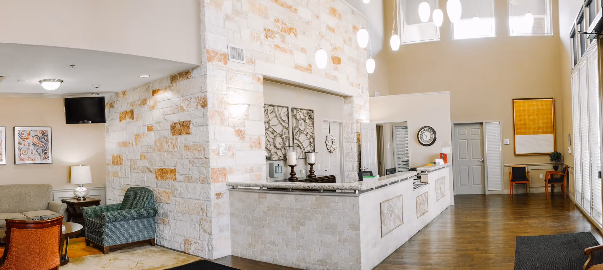 A spacious and well-lit reception area with a stone accent wall behind the front desk. The desk has a granite countertop and decorative panels. To the left, there is a seating area with a green armchair, an orange chair, a beige sofa, and a side table with a lamp. The walls are adorned with framed artwork and a clock. Large windows on the right side allow natural light to fill the room, and modern pendant lights hang from the ceiling.