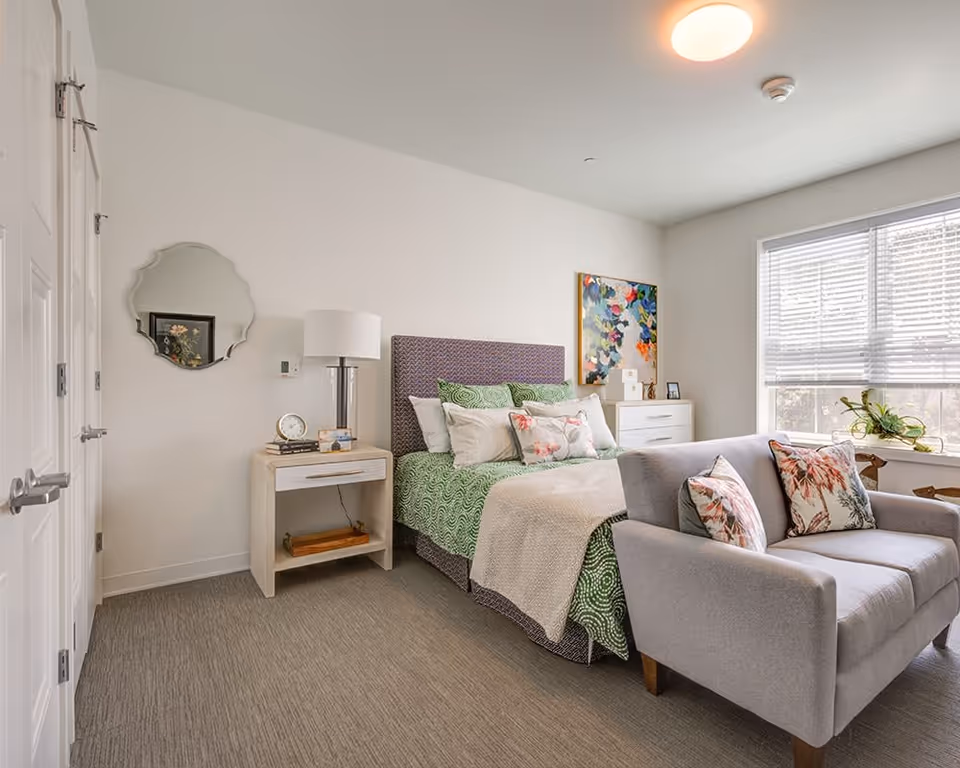 A bright and cozy bedroom featuring a bed with green and white bedding, multiple pillows, and a gray upholstered headboard. In front of the bed is a light gray loveseat with floral pillows. To the left of the bed is a nightstand with a lamp, clock, and books. A decorative mirror hangs on the wall above the nightstand. On the right side, there is a white dresser with a colorful abstract painting above it and a large window with blinds letting in natural light. The room has light-colored walls and carpeted flooring.