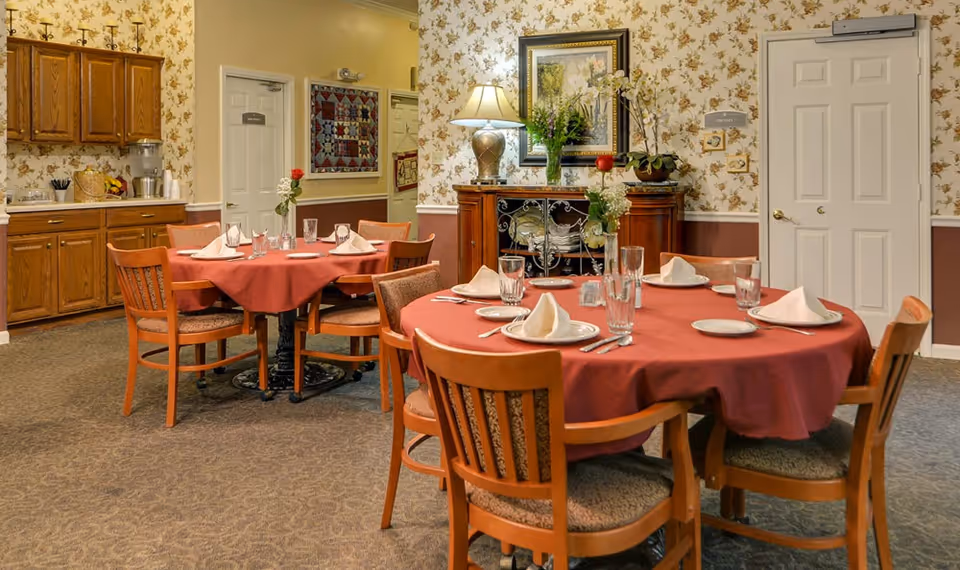 Dining room in a senior living facility with round tables covered in red tablecloths, set with plates, glasses, napkins, and silverware. Wooden chairs surround the tables. The room has floral wallpaper, a sideboard with a lamp and flowers, and wooden cabinets along one wall.