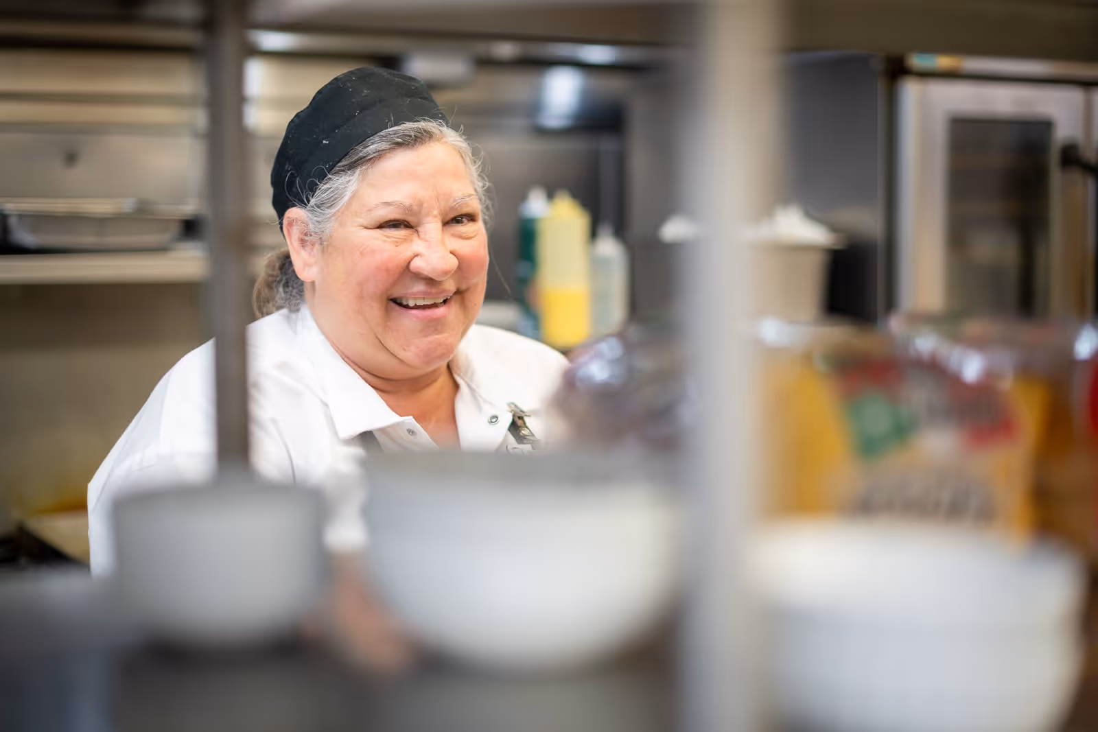 A smiling woman wearing a white chef's coat and a black hairnet is seen in a kitchen setting with blurred kitchen equipment and containers in the foreground and background.