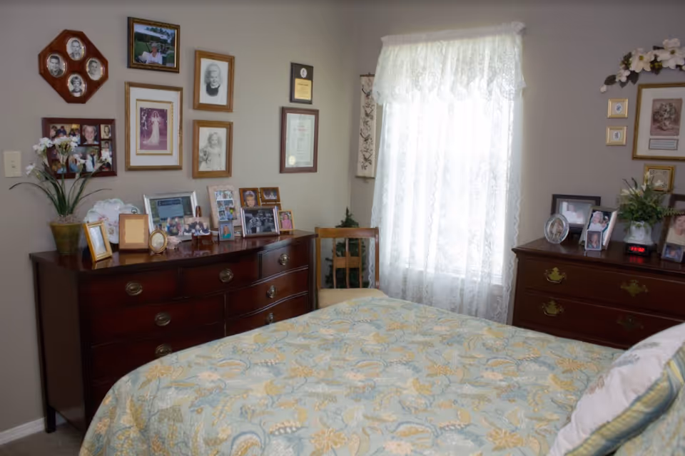 Cozy bedroom with a floral bedspread, two wooden dressers covered in framed photos, wall art, and a lace-curtained window.