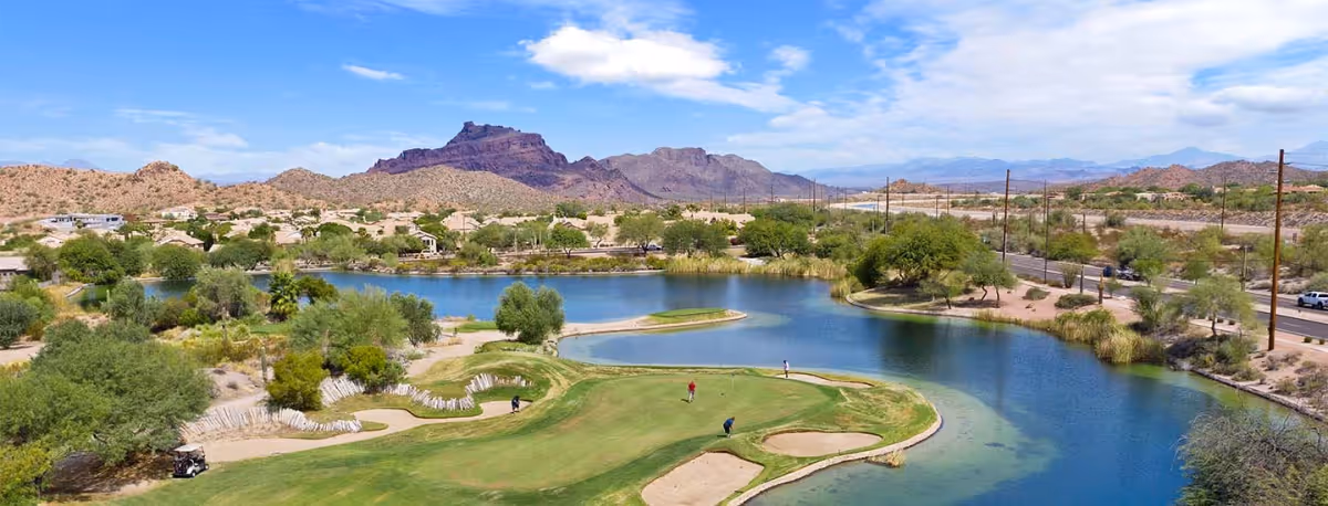 A scenic view of a golf course with a water hazard surrounded by desert landscape and mountains in the background under a partly cloudy blue sky. There are a few people playing golf on the green.