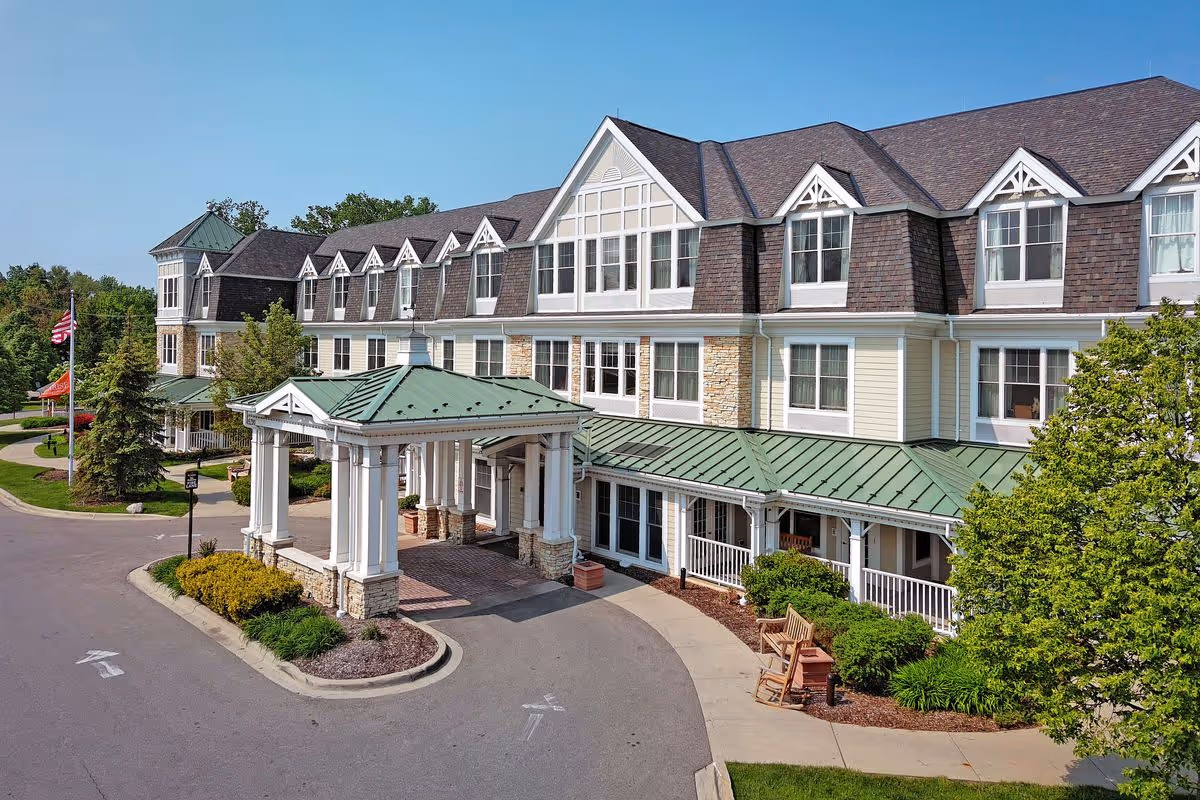 Exterior view of a large senior living facility building with a covered entrance, multiple windows, and a green metal roof. The building is surrounded by landscaped greenery, benches, and a driveway. The sky is clear and blue.