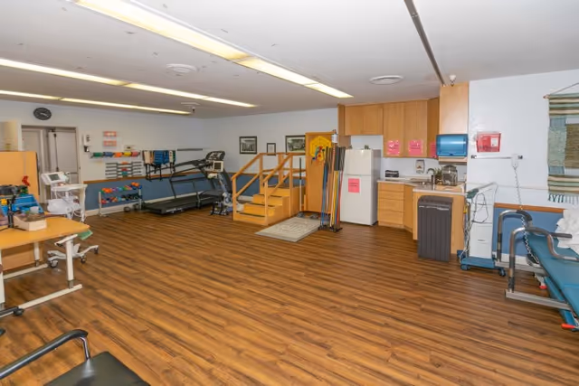 Large therapy room with treadmills, exercise equipment, parallel bars, a kitchenette area, and wood-patterned flooring.