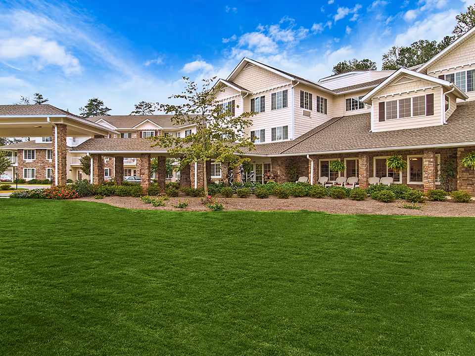 Front exterior of a multi-story senior living building with a covered entrance, landscaped lawn, and porch seating.