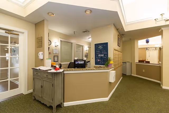 Reception area inside Granville Place senior living facility with a front desk, a small cabinet with papers and a red bow, a chalkboard with a welcoming message, mailboxes on the wall, and a hallway leading to other rooms.