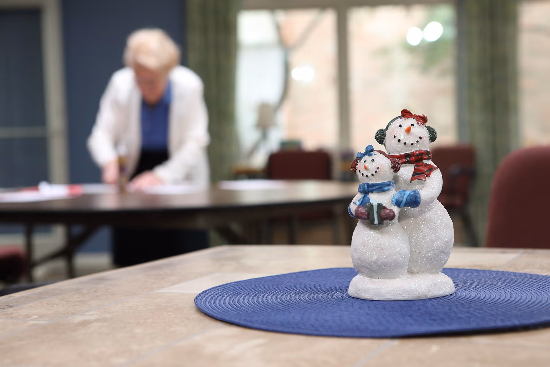A small decorative snowman figurine with a red scarf and earmuffs stands on a blue placemat on a table in a room. In the background, an elderly person is slightly blurred, standing near another table with papers.
