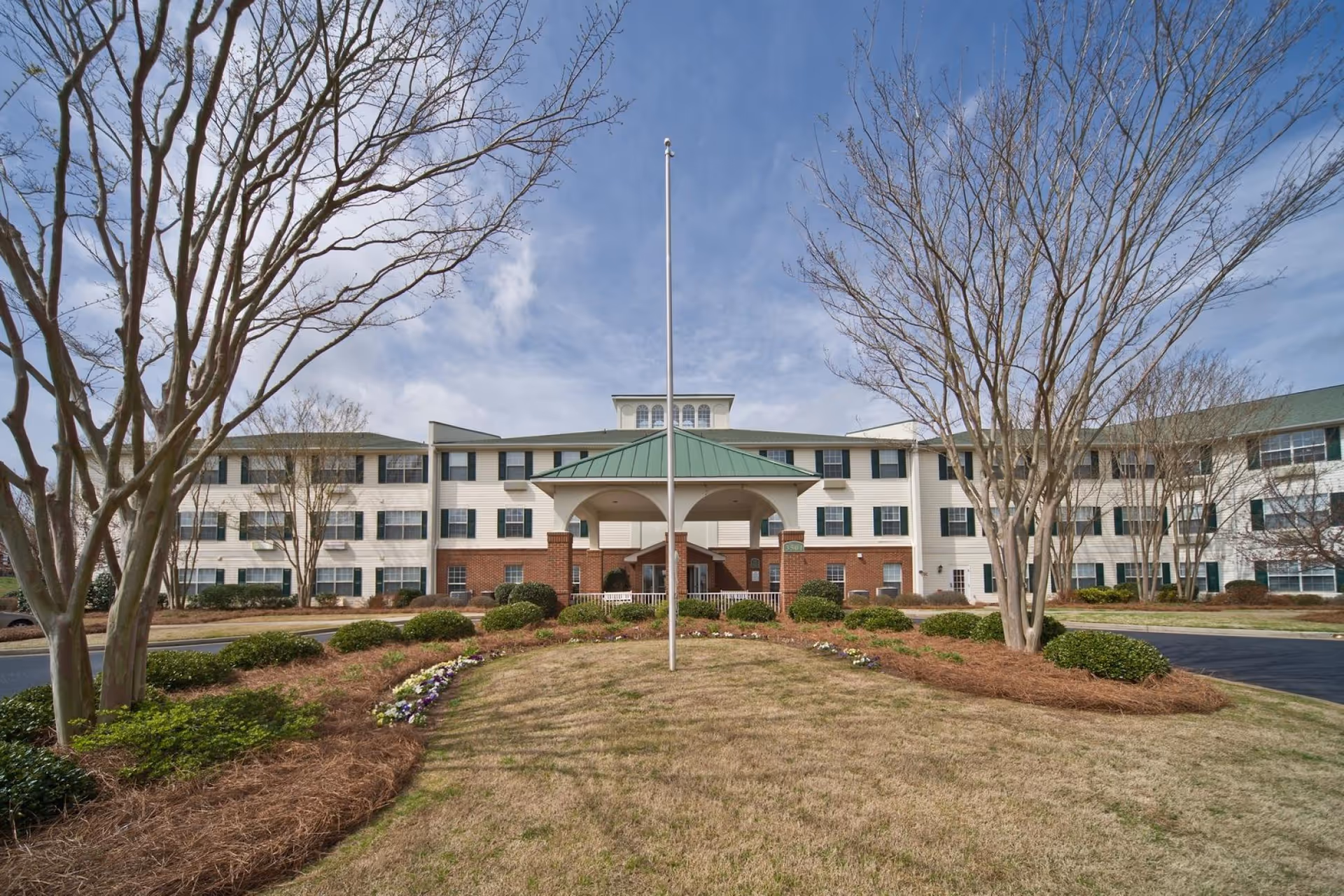 Front exterior view of The Legacy of Anderson senior living facility with a circular driveway, landscaped garden with bushes and flowers, leafless trees, and a flagpole in the center under a partly cloudy sky.