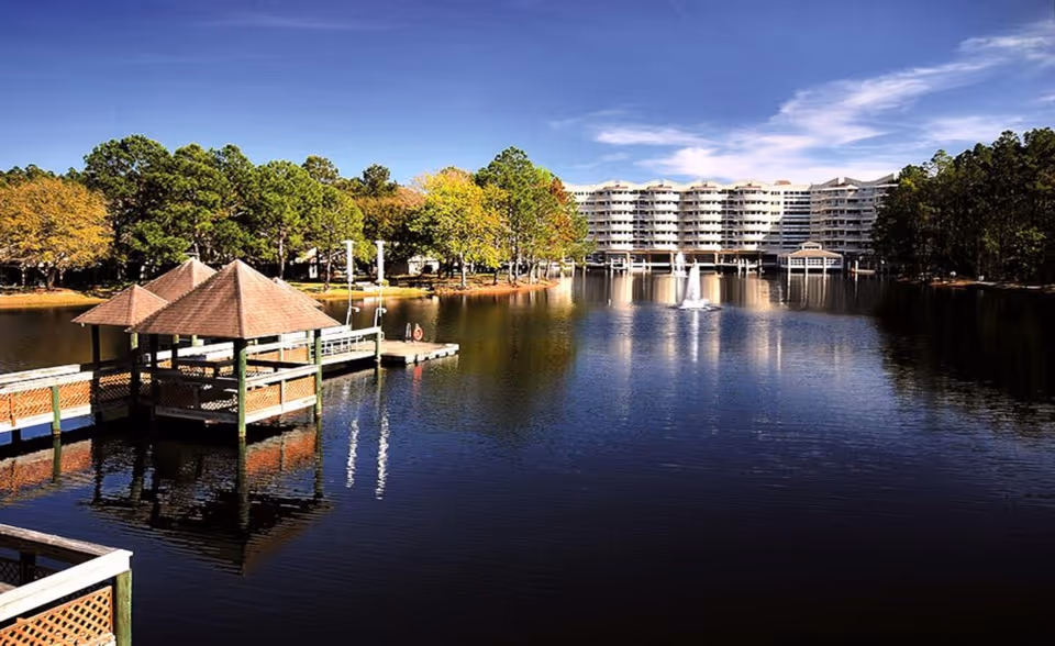View across a lake with a gazebo and dock in the foreground and a multi-story residential building with fountains in the background.