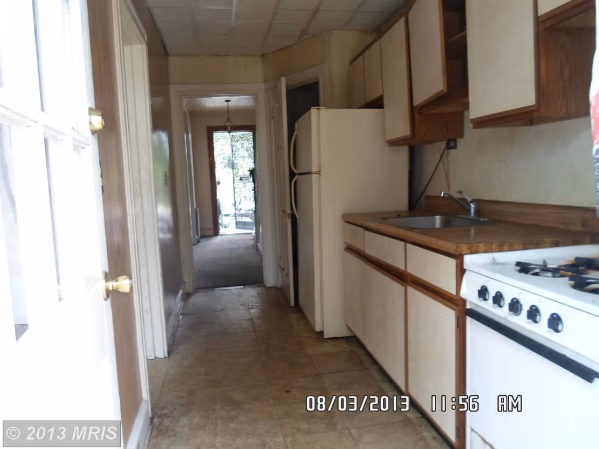 A narrow kitchen with a white refrigerator, wooden cabinets, a countertop with a sink, and a white gas stove. The floor is tiled, and there is a hallway leading to another room with a door and window letting in natural light.