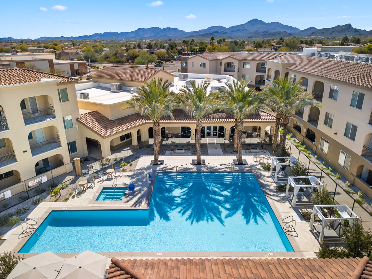 Aerial view of a senior living facility's outdoor pool area with a large rectangular swimming pool, a smaller hot tub, several palm trees casting shadows on the water, lounge chairs, and cabanas. Surrounding the pool are multi-story buildings with balconies, and mountains are visible in the background under a clear blue sky.