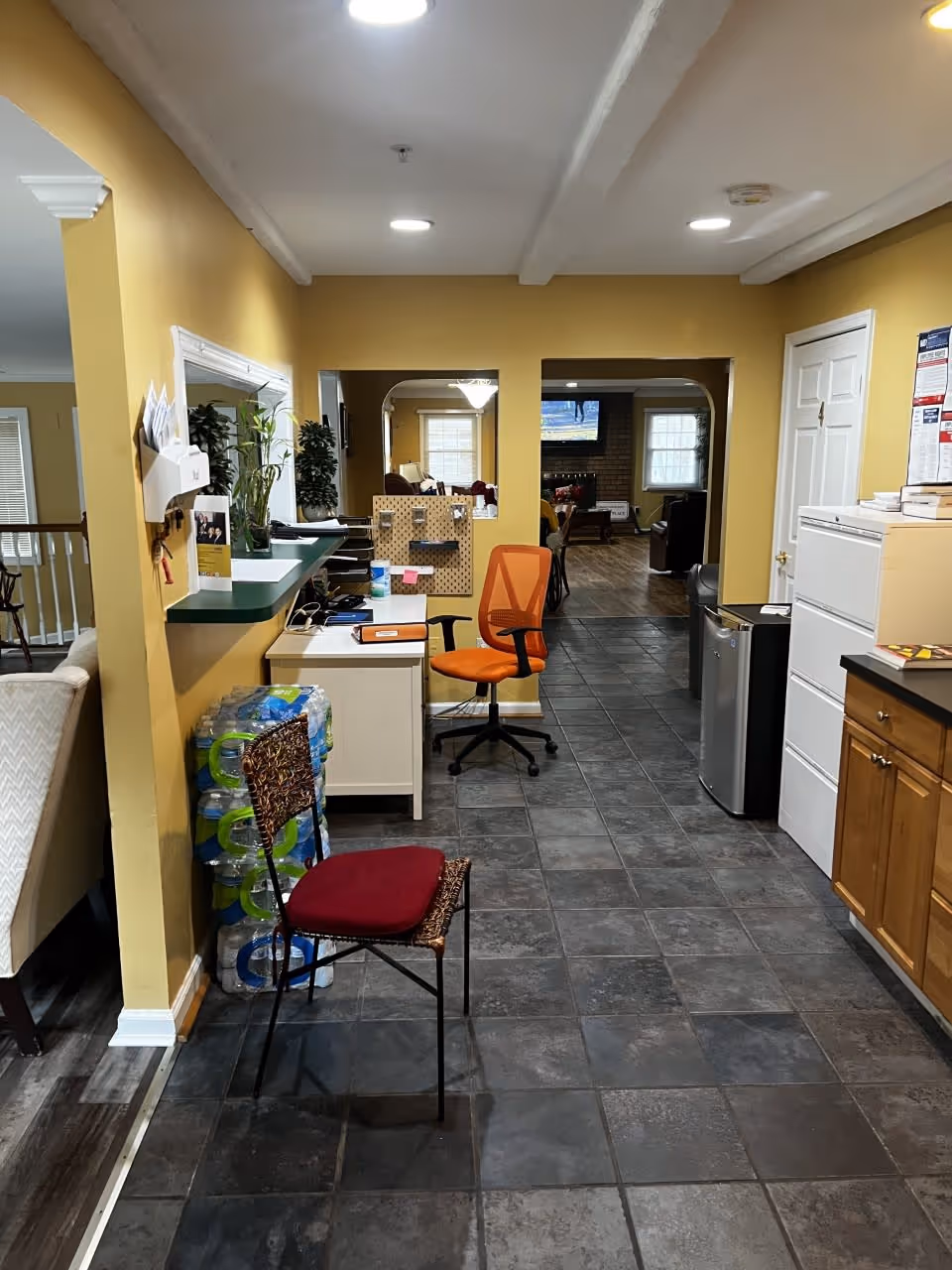 Interior view of a facility hallway with yellow walls and dark tiled floor. There is a small desk with an orange office chair and a red cushioned chair in the foreground. Water bottles are stacked against the wall near the desk. The hallway leads to a living area with windows and a TV mounted on the wall. Cabinets and a mini fridge are visible on the right side.