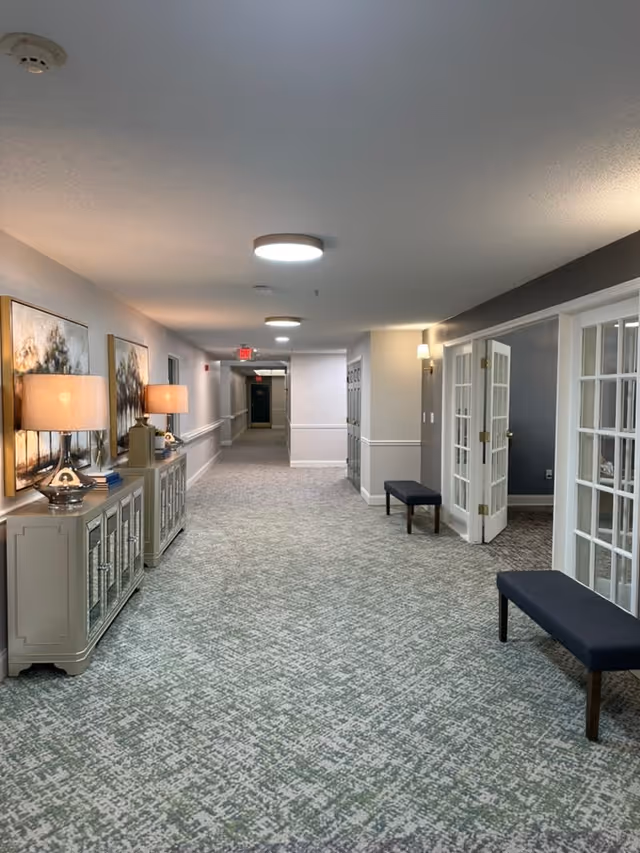 A well-lit hallway in an assisted living facility with patterned carpet flooring, light gray walls, and ceiling lights. On the left side, there are two decorative cabinets with table lamps and framed artwork above them. On the right side, there are two benches and a set of open French doors leading to another room.