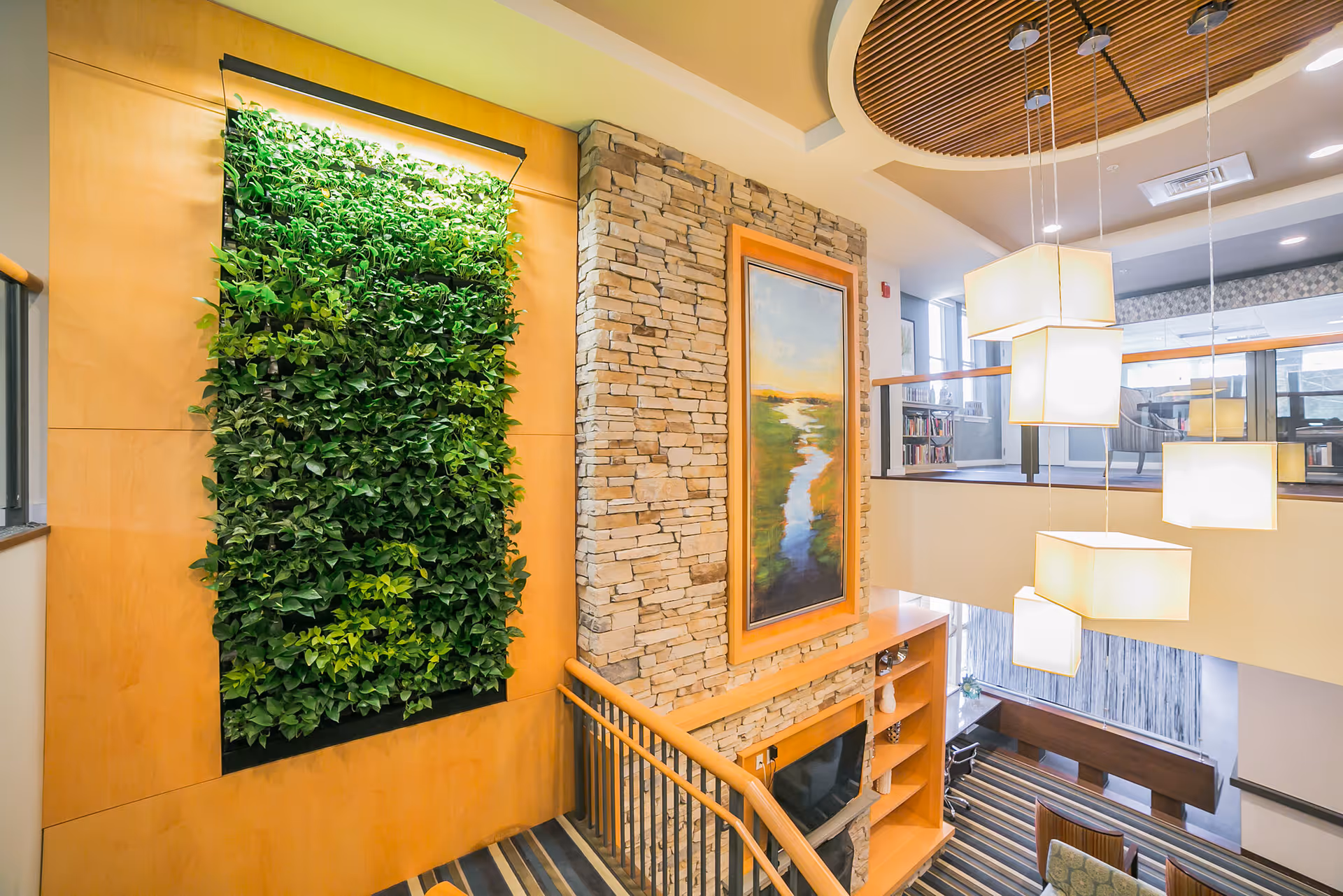 Interior view of a senior living facility featuring a vertical green plant wall, a stone accent wall with a framed landscape painting, a wooden staircase railing, modern hanging square pendant lights, and a seating area with striped carpet below.