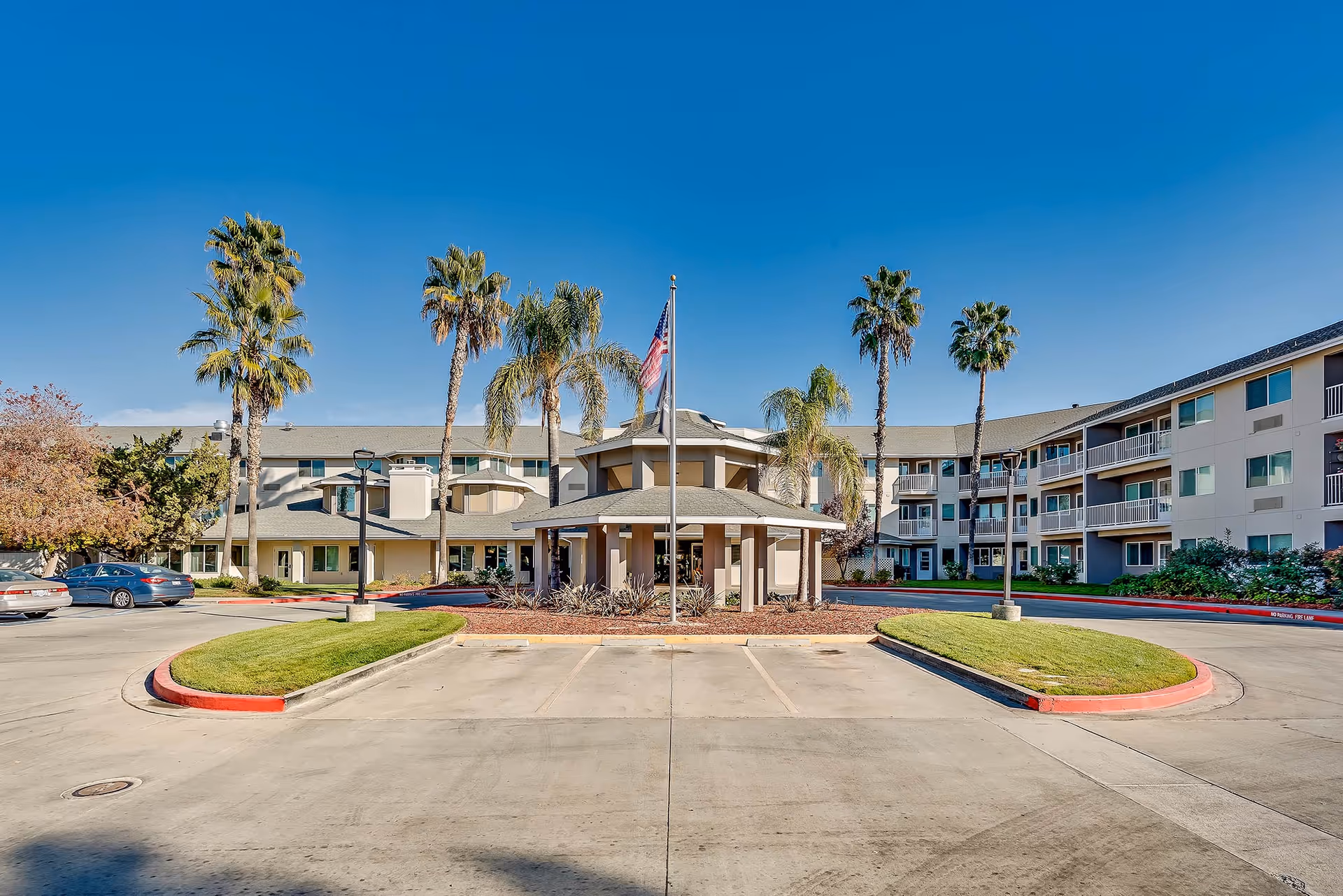 Exterior view of The Remington Senior Living facility showing a three-story building with balconies, palm trees, an American flag on a flagpole, and a circular driveway with parked cars under a clear blue sky.