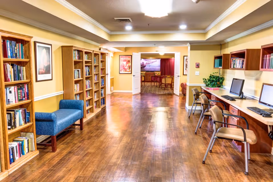 A well-lit interior hallway with wooden floors and yellow walls. On the left side, there are multiple wooden bookshelves filled with books and a blue upholstered bench. On the right side, there is a long desk with three chairs and two computer monitors. At the end of the hallway, double doors open into another room with chairs and a large screen or picture on the wall.