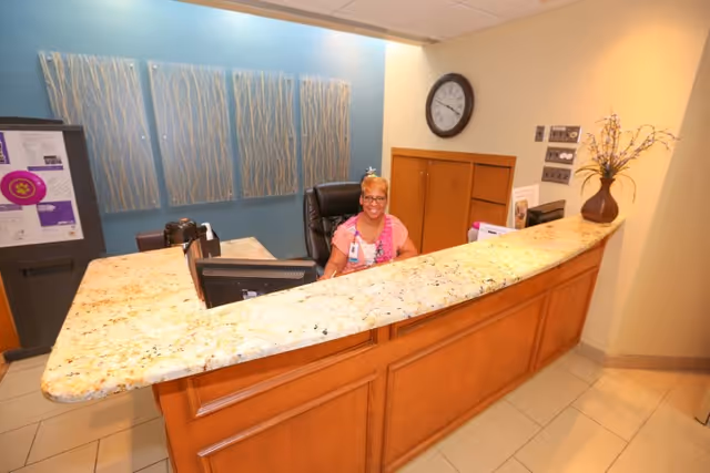 Reception area with a curved granite countertop desk, a smiling receptionist seated behind the desk, a wall clock, decorative wall art, and a vase with flowers on the counter.