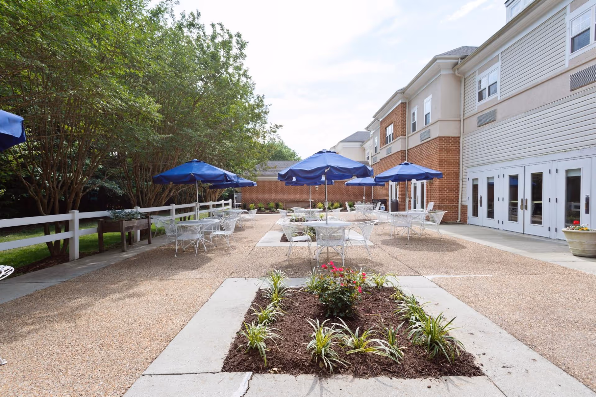 Outdoor courtyard patio with white metal tables and blue umbrellas, a planted flower bed in the foreground, and the facility building with doors on the right.