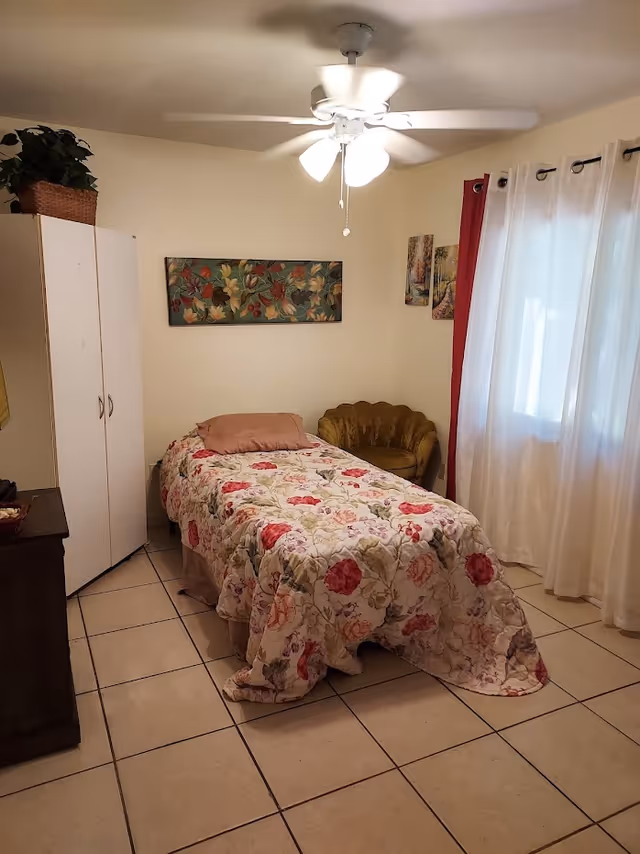 A small bedroom with a single bed covered in a floral quilt, a brown pillow, a white wardrobe with a plant on top, a brown cushioned chair, three pieces of wall art, a ceiling fan with lights, and a window with white curtains and red drapes.