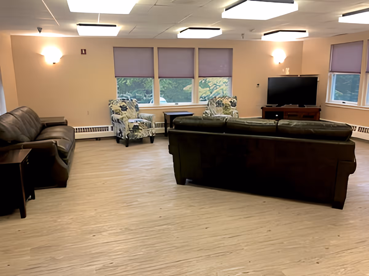 A spacious living room with beige walls and light wood flooring. The room features two floral-patterned armchairs near three windows with purple blinds, a dark brown leather sofa facing away from the camera, another dark brown leather sofa on the left side, a wooden side table, and a flat-screen TV on a wooden stand in the corner.