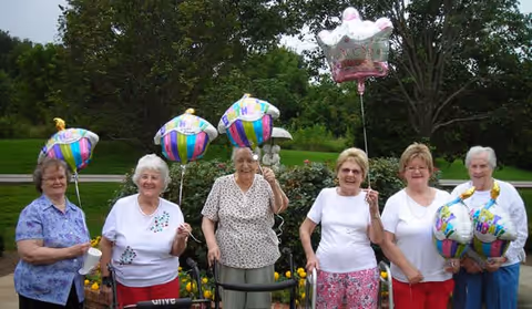 Six elderly women standing outdoors in a garden area, each holding colorful birthday balloons. They are smiling and appear to be celebrating a birthday together, with greenery and trees in the background.