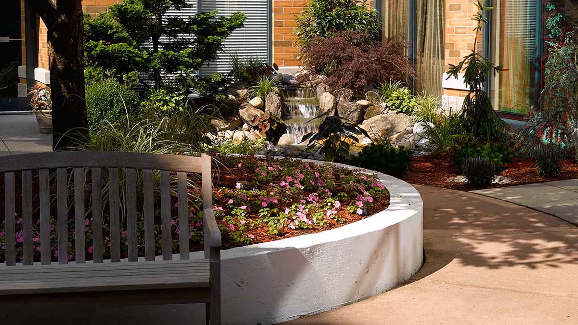 Outdoor garden area at Atria Cutter Mill featuring a wooden bench, a curved white planter filled with pink flowers and greenery, a small waterfall cascading over rocks, and surrounding plants and trees near a brick building.