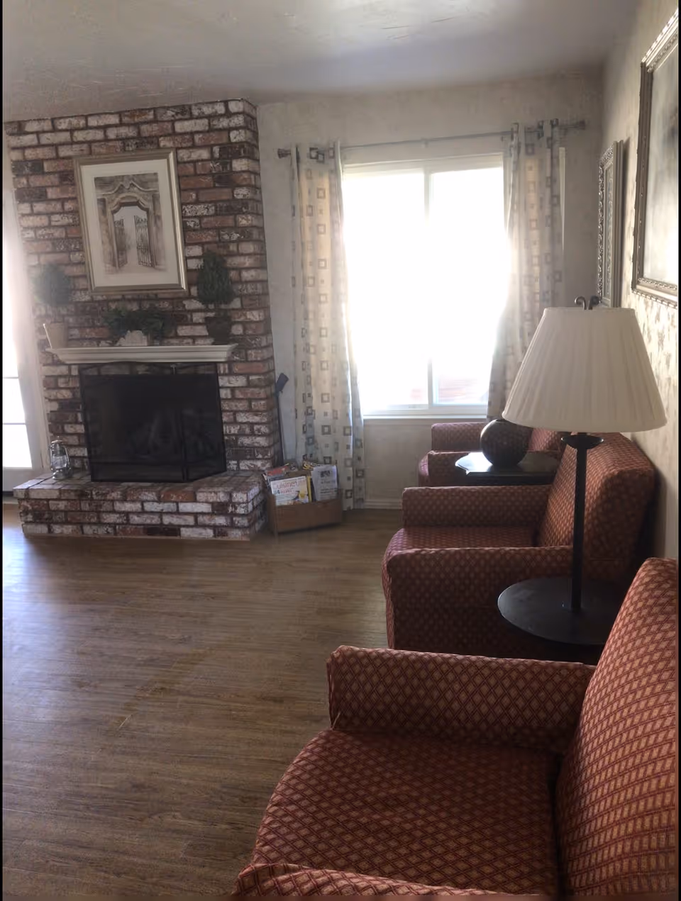 Sunlit living room with patterned armchairs and side tables, a floor lamp, a brick fireplace, and a window with curtains.