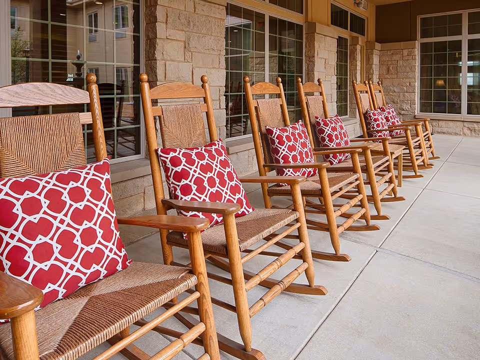 A row of wooden rocking chairs with red and white patterned cushions lined up on a covered patio outside a building with stone walls and large windows.