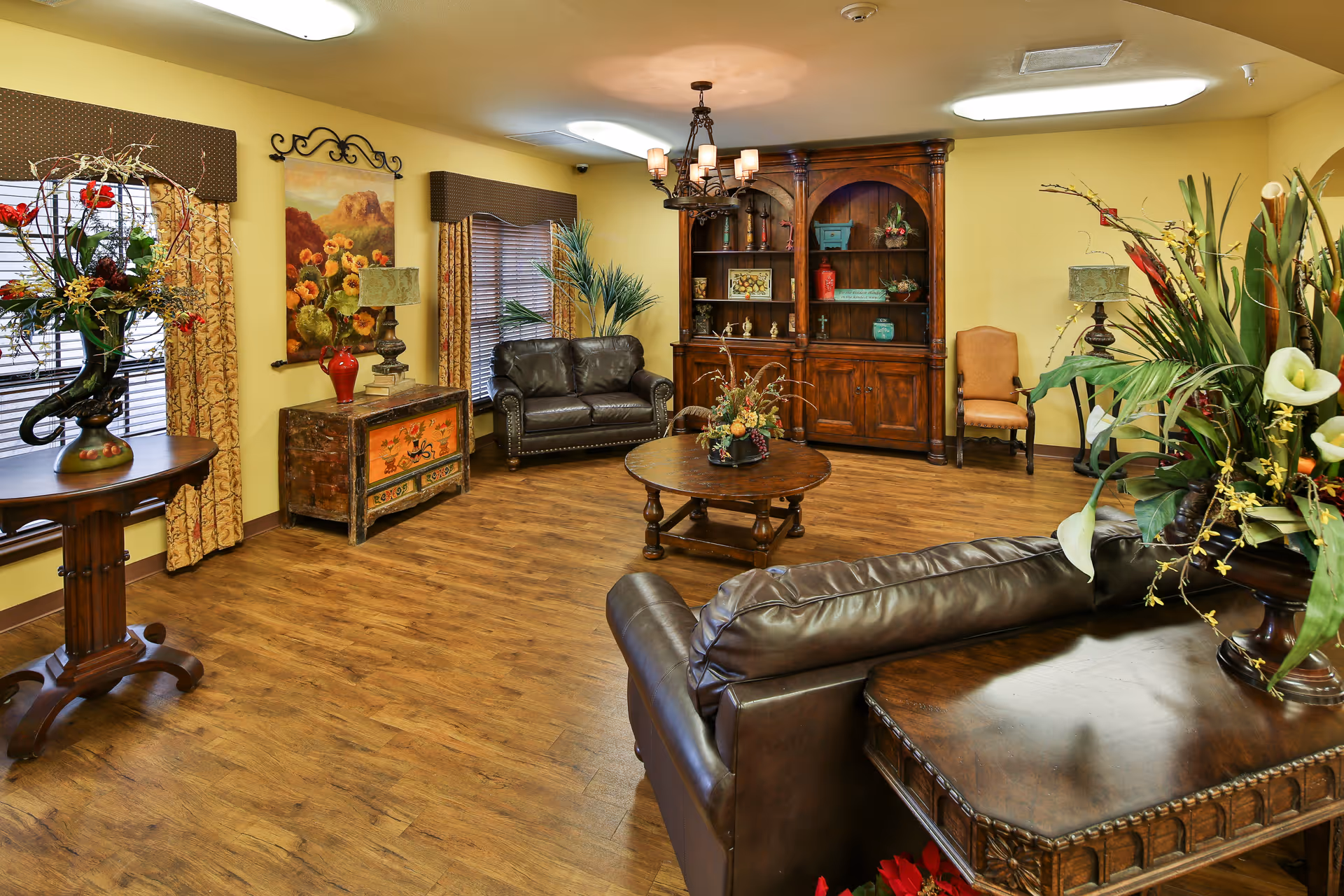 A cozy living room with wooden flooring, yellow walls, and multiple pieces of dark wood furniture including a large cabinet, coffee table, and side tables. There are two leather sofas, a single chair, decorative lamps, floral arrangements, and framed artwork on the walls. The room is warmly lit with ceiling lights and a chandelier.
