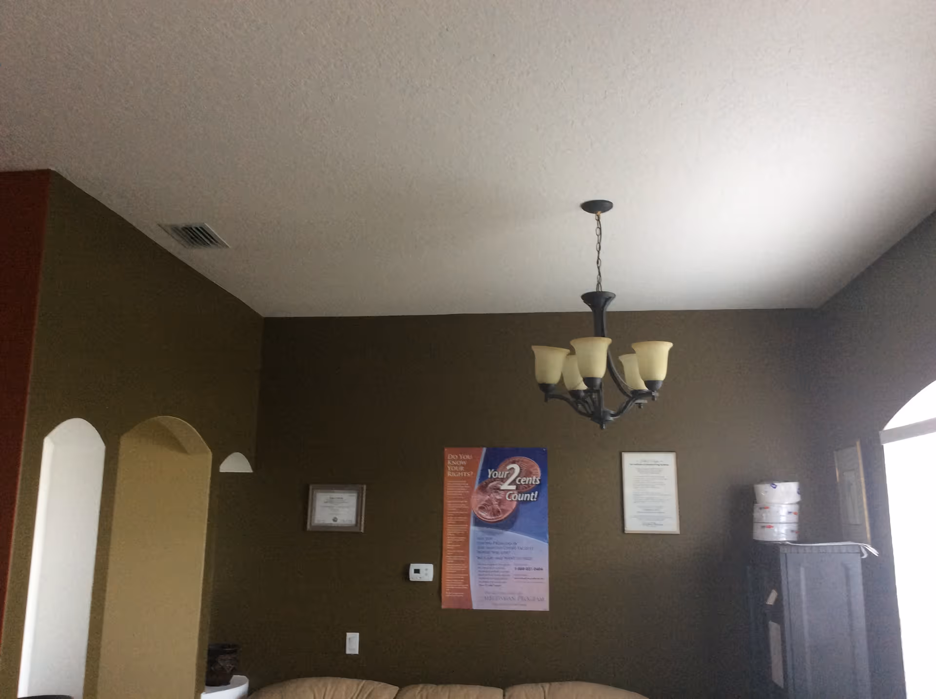 Interior view of a room with olive green walls and a beige ceiling. A chandelier with five frosted glass shades hangs from the ceiling. On the wall, there are three framed items including a colorful poster in the center. A beige couch is partially visible at the bottom of the image. There is an arched doorway on the left and a window on the right with some stacked items near it.