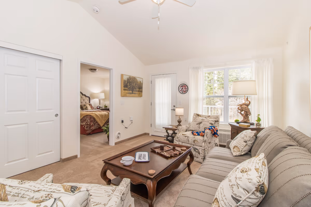 Bright living room with a striped sofa, patterned armchairs, wooden coffee table, and a doorway leading to a bedroom and exterior door.