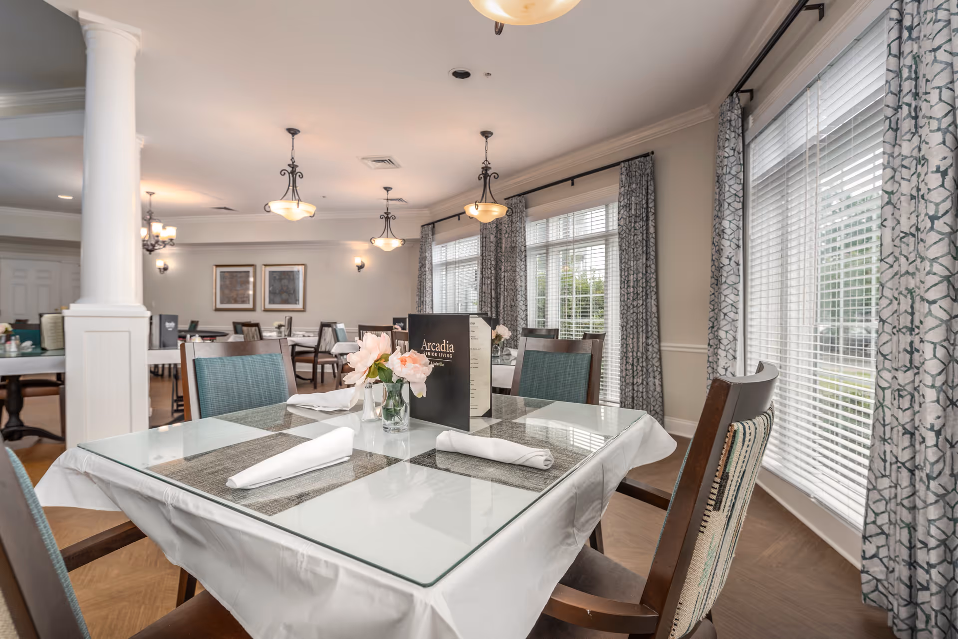 A bright dining room with large windows covered by patterned curtains. The tables are set with white tablecloths, glass tops, neatly folded white napkins, and small flower arrangements. A menu labeled 'Arcadia Senior Living' is placed on the table in the foreground. The room features elegant light fixtures and framed artwork on the walls.