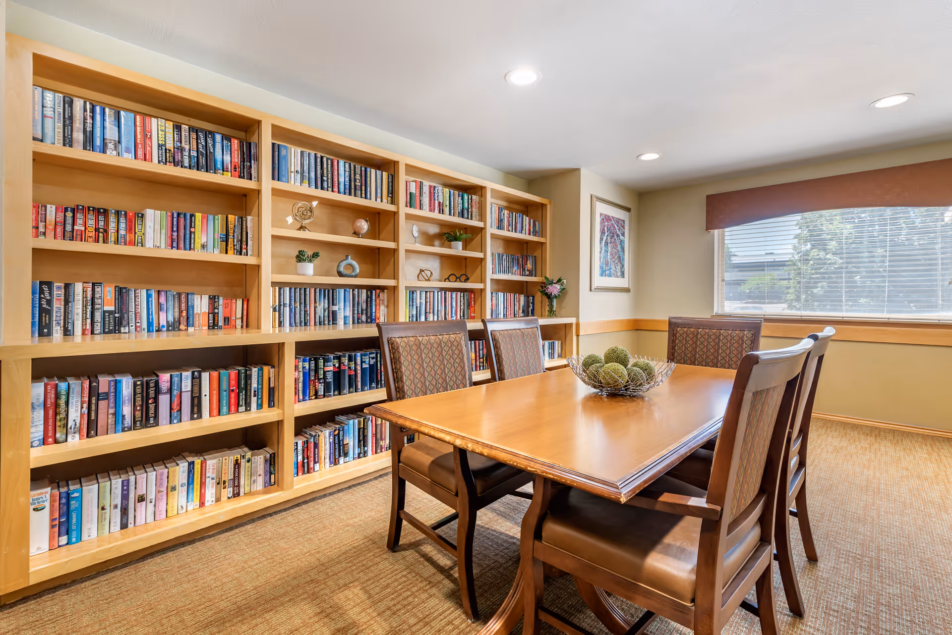 A cozy communal reading room with built-in bookshelves, a wooden table and chairs, and a window with blinds.