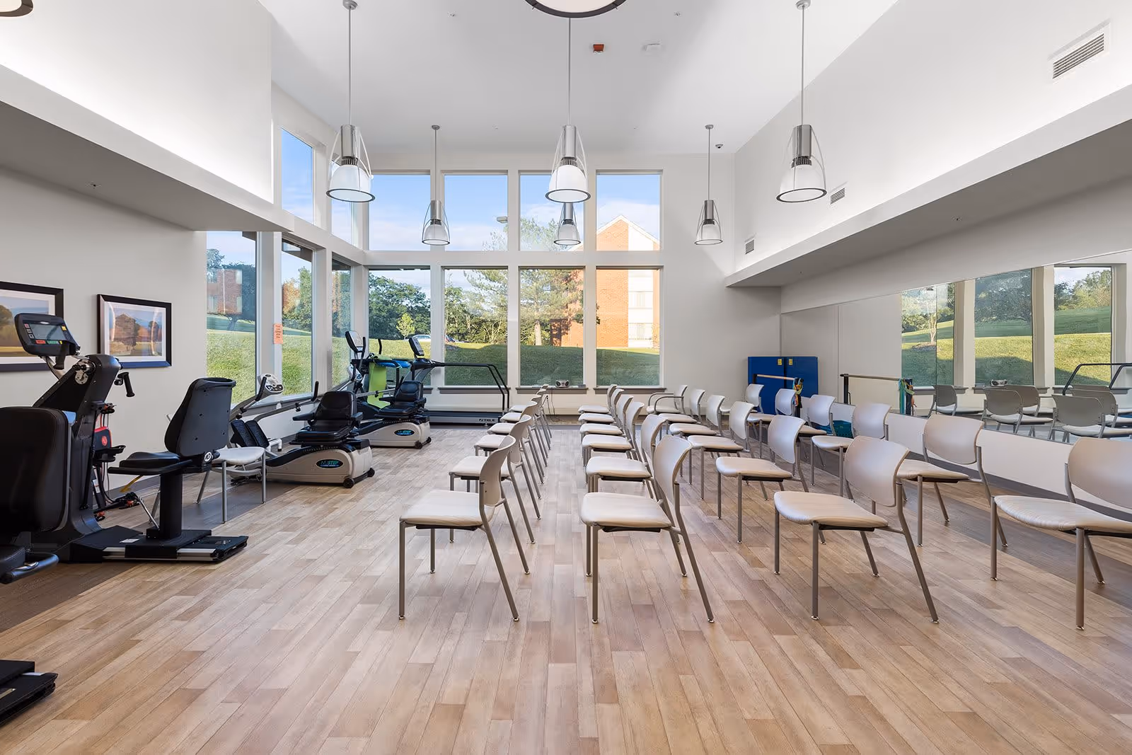 A bright and spacious exercise room with large windows letting in natural light. The room features several exercise machines on the left side and rows of chairs arranged in the center and right side. The walls are white, and there are pendant lights hanging from the ceiling. A large mirror covers the right wall, reflecting the room and the outdoor greenery visible through the windows.