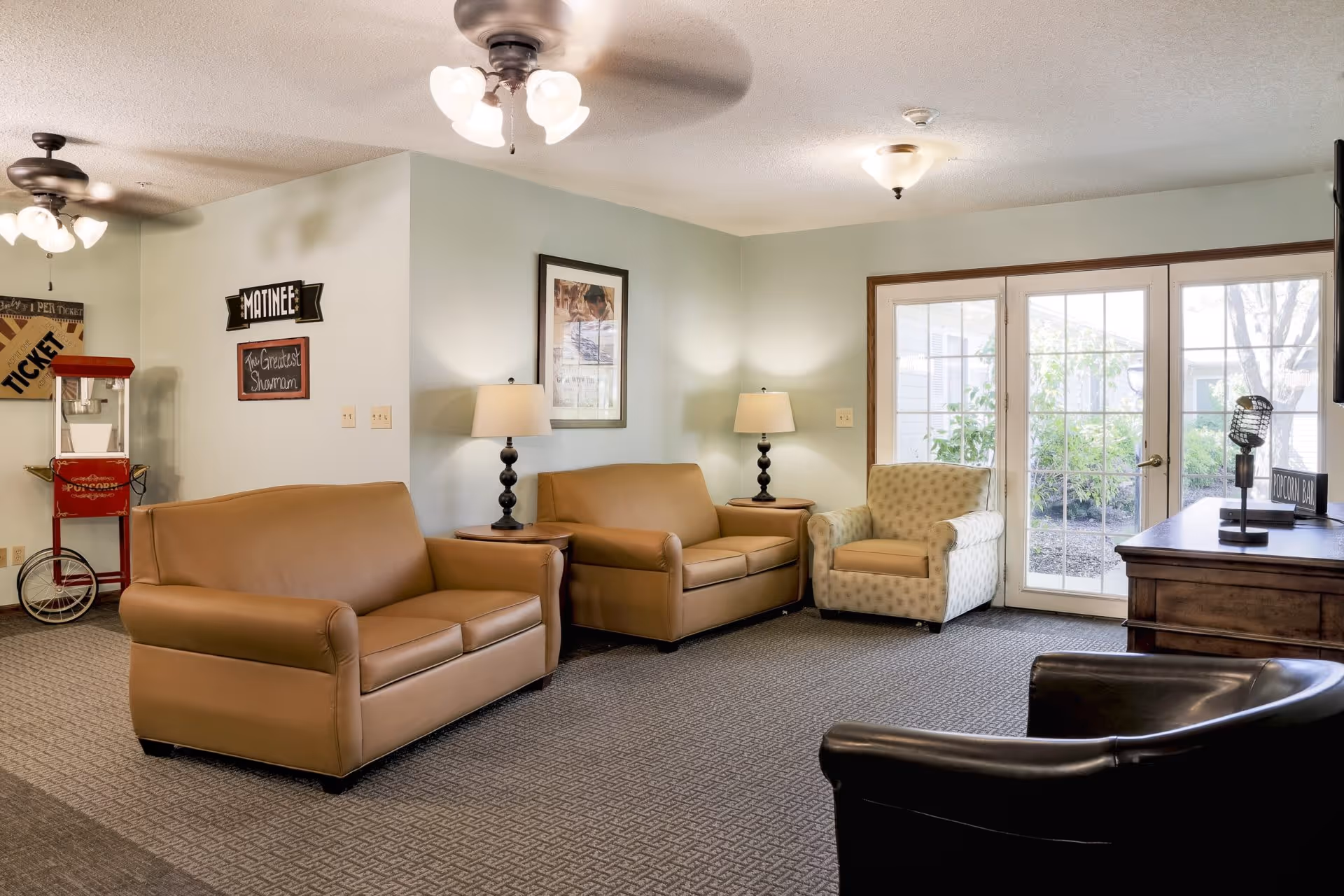A cozy living room area with two brown leather loveseats, a patterned armchair, and a black leather chair. The room has light green walls, two table lamps on wooden side tables, ceiling fans with lights, and a popcorn machine in the corner. French doors with glass panes lead outside to a garden area.