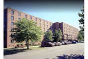 Exterior view of a multi-story brick building with several windows, a parking lot with cars, and trees along the sidewalk under a clear sky.