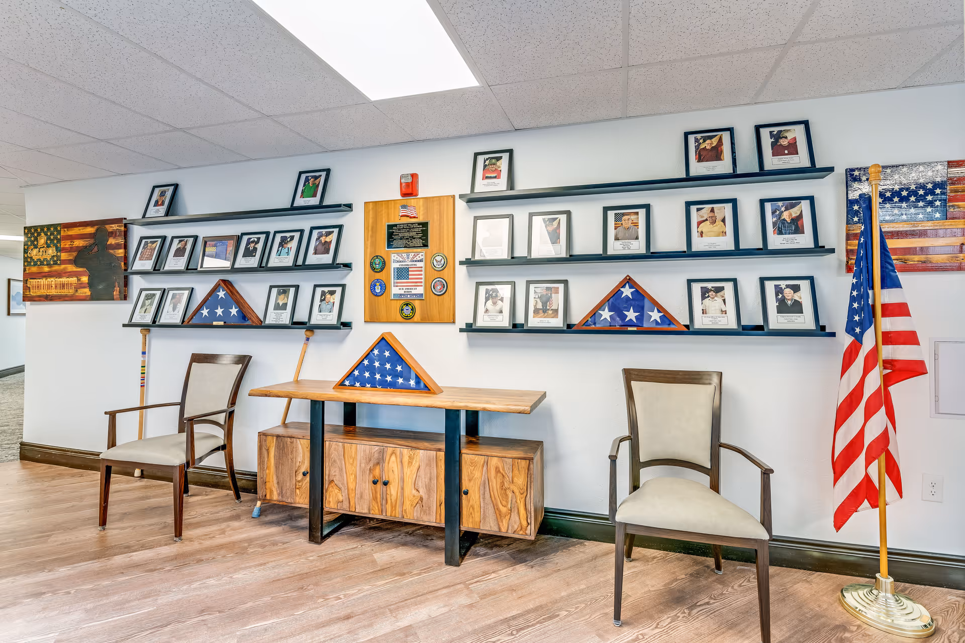 Interior wall display in a senior living facility featuring framed photographs of veterans, folded American flags in triangular cases, military insignia plaques, two chairs, a wooden table, and an American flag on a stand.