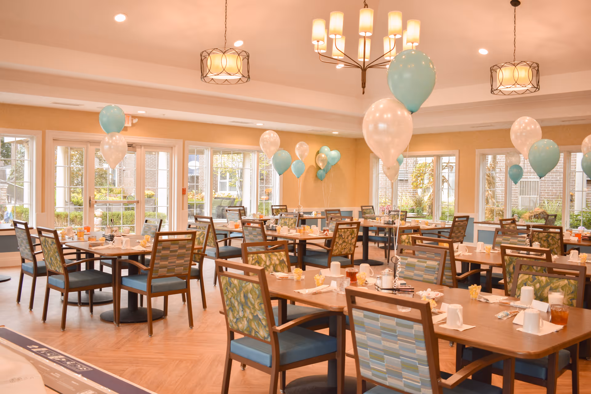 A bright dining room decorated with teal and white balloons tied to the backs of chairs. The room has multiple wooden tables with chairs that have patterned upholstery. Each table is set with cups, napkins, and utensils. Large windows allow natural light to fill the space, and pendant lights hang from the ceiling.