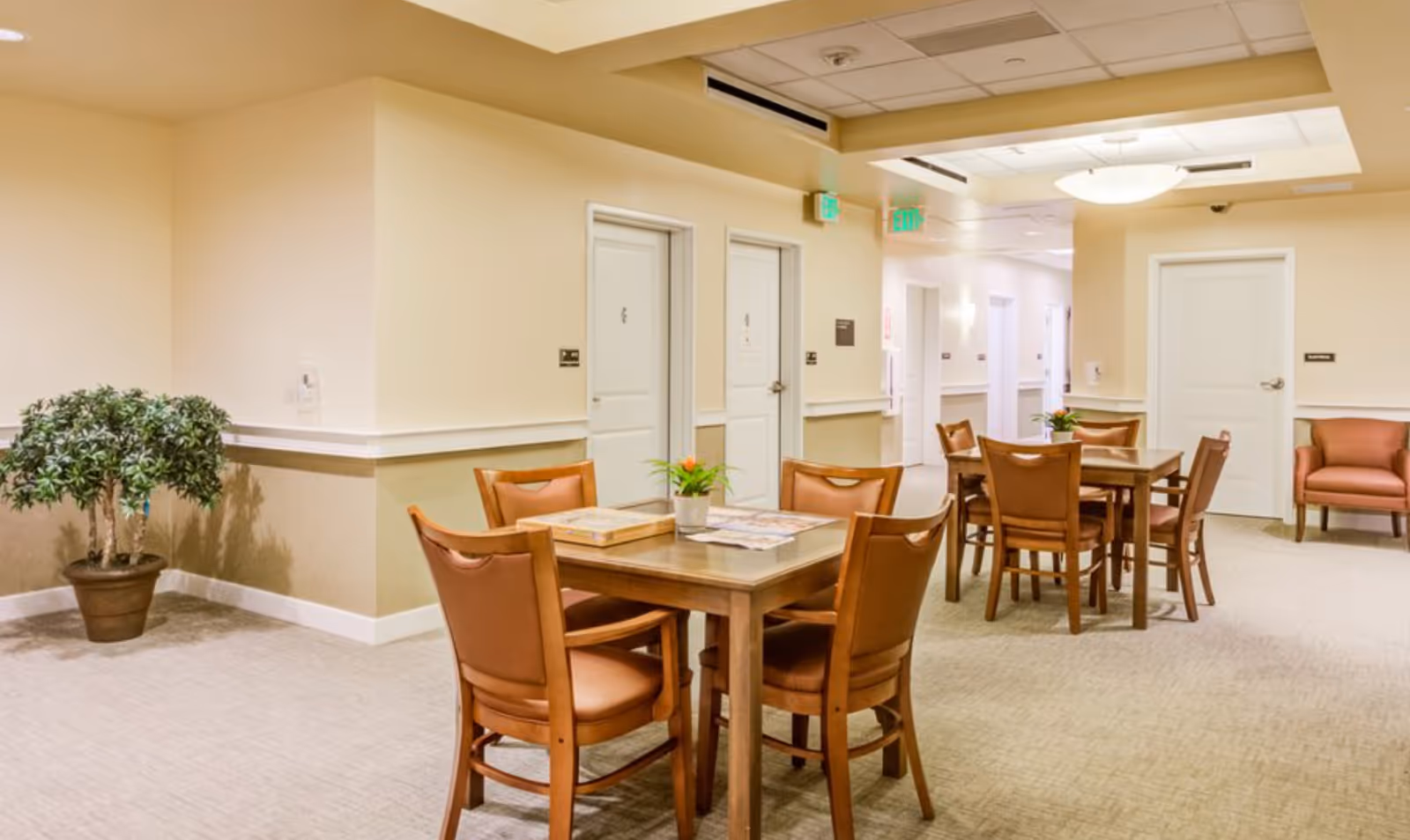 A well-lit interior common area with two wooden tables, each surrounded by four brown cushioned chairs. There are small potted plants on the tables and a potted plant in the corner. The walls are painted beige with white trim, and there are several white doors and exit signs visible in the background.