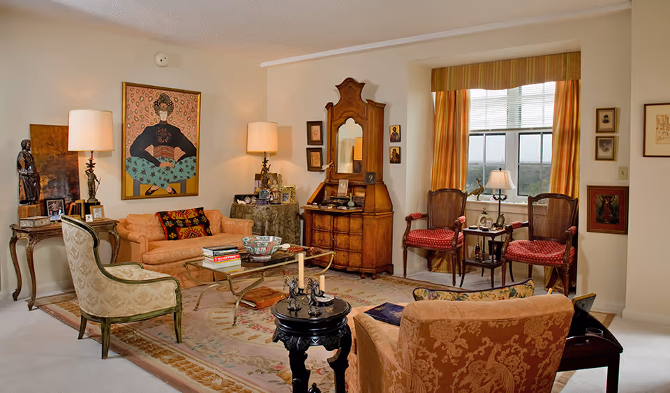 A warmly decorated living room with upholstered sofas and chairs, wooden cabinets and tables, artwork on the walls, and a window with drapes.