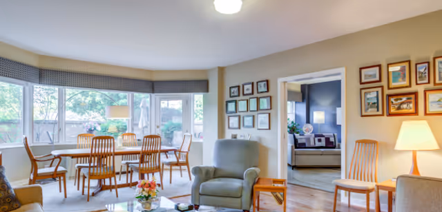 A bright living room with large windows overlooking a garden. The room features a dining table with six wooden chairs, a comfortable armchair, a glass coffee table with a flower arrangement, and framed pictures on the walls. There is a doorway leading to another room with a sofa and lamp.